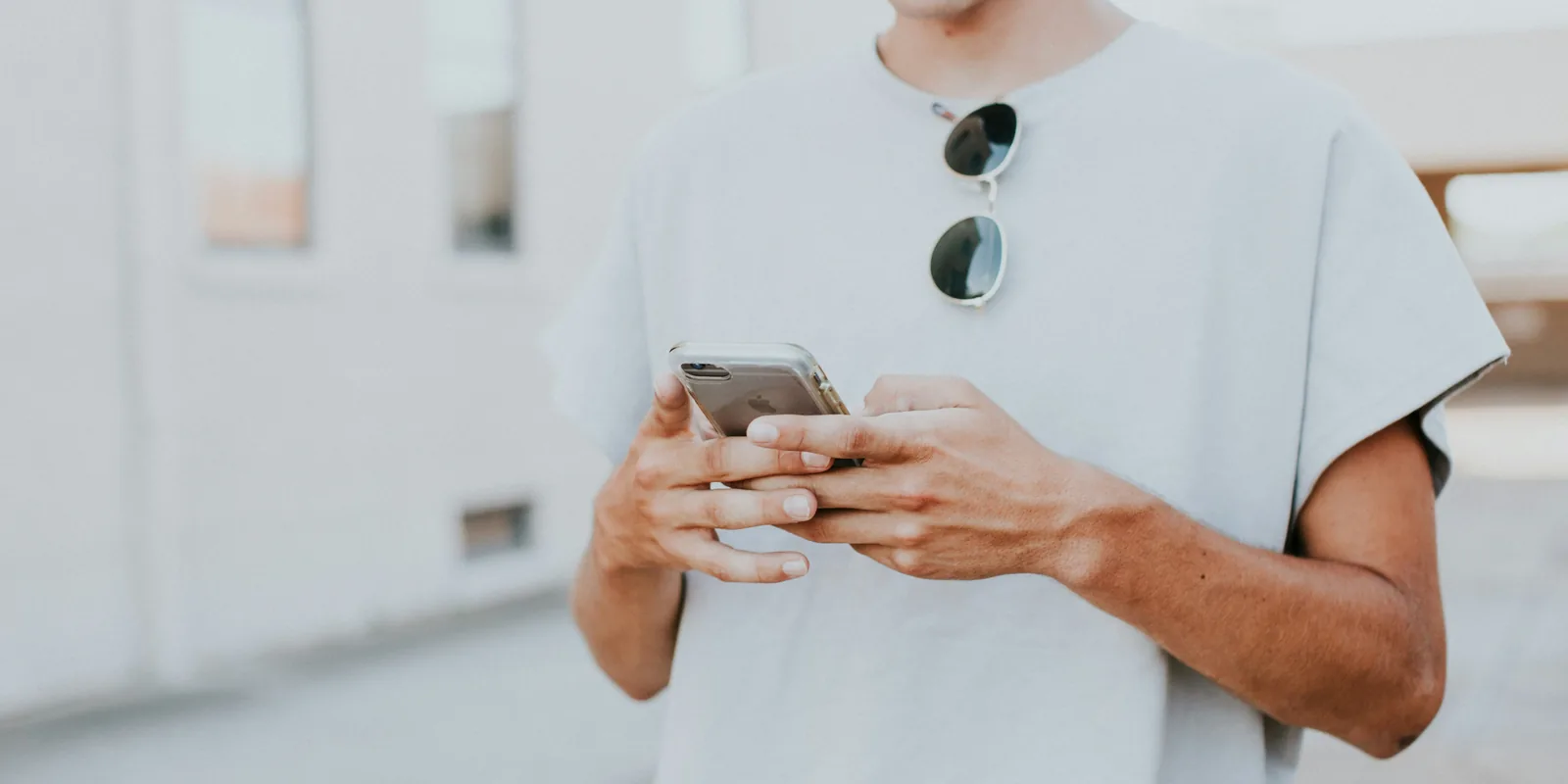 Eine Person der Gen Z mit weißem T-Shirt, Sonnenbrille und Handy vor der Brust ist zu sehen. Sie schreibt eine SMS